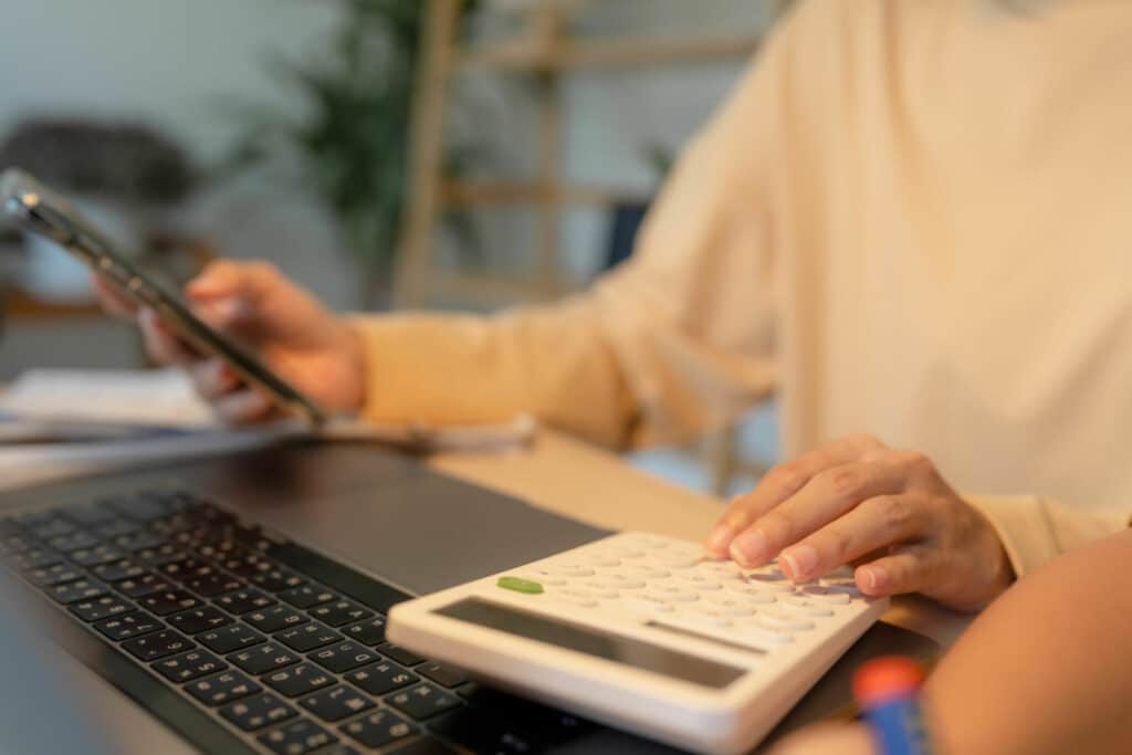 Young Asian mother looking at smart phone and using calculator while working on laptop next to her son who is playing crayon drawing.